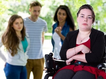 A young woman in a wheelchair with her arms crossed. Behind her is a group of people who are talking about her